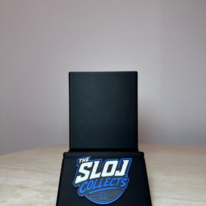 May include: A black display stand with the text "THE SLOJ COLLECTS" in white and blue. The stand is rectangular with a flat base and a vertical back. The stand is designed to hold a trading card.