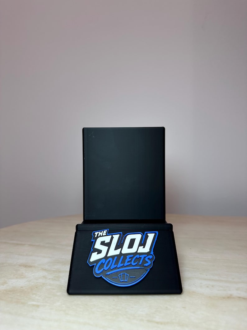 May include: A black display stand with the text "THE SLOJ COLLECTS" in white and blue. The stand is rectangular with a flat base and a vertical back. The stand is designed to hold a trading card.