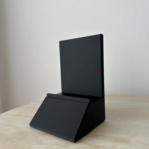 May include: A black wooden display stand with a rectangular back and a slanted base. The stand has a small ledge at the bottom of the back piece. The stand is sitting on a light-colored surface.