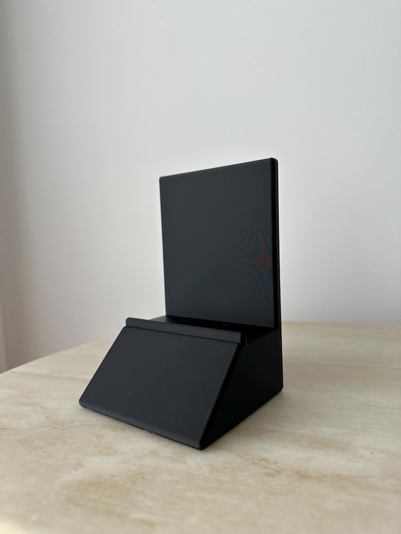 May include: A black wooden display stand with a rectangular back and a slanted base. The stand has a small ledge at the bottom of the back piece. The stand is sitting on a light-colored surface.