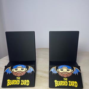 May include: Two black display stands with a rectangular shape. Each stand features a graphic of a bearded cartoon character with blue wings and a blue cap. The text "THE BEARDED ZARD" is printed in yellow.