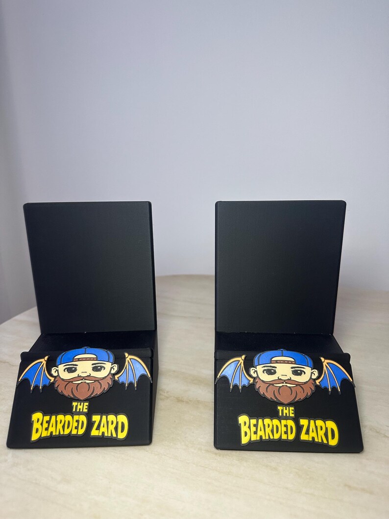 May include: Two black display stands with a rectangular shape. Each stand features a graphic of a bearded cartoon character with blue wings and a blue cap. The text "THE BEARDED ZARD" is printed in yellow.
