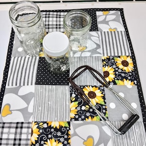 May include: A rectangular quilted table runner featuring a patchwork pattern of sunflowers, bees, black and white plaid, stripes, and polka dots. Three glass mason jars and a metal jar lifter with black handles are placed on the fabric surface.