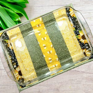 May include: A clear glass baking dish rests on a colorful quilted pad. The pad features vertical stripes of yellow with bee patterns, olive green, and black with sunflower designs. The dish has a rectangular shape with rounded corners and handles.