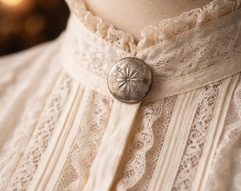 May include: A close-up of a cream-coloured vintage blouse with detailed lace. The blouse has a high, frilled collar and a decorative silver button with a starburst design. The fabric appears delicate, and the style is reminiscent of Victorian era clothing.