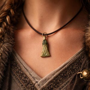Puede incluir: Collar con colgante de bronce y cordón negro. El colgante tiene forma triangular con líneas grabadas y un pequeño lazo sujeto a un anillo dorado. El collar se lleva sobre una prenda con ribete de piel.