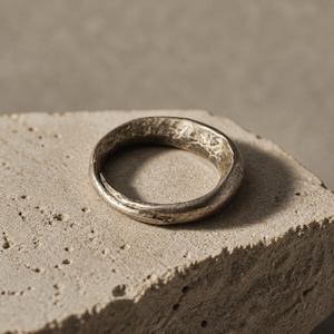 May include: A silver-toned ring with a textured, uneven surface and a rounded band. The metal has a matte, weathered finish with visible indentations and a slightly irregular, organic shape that gives it a distinct, non-uniform appearance.