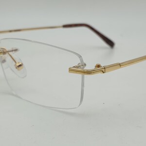 May include: Rimless spectacles featuring clear rectangular lenses and a polished gold-tone metal frame. The thin temples transition into dark brown acetate tips, and the bridge connects the lenses with gold-tone hardware and adjustable nose pads.