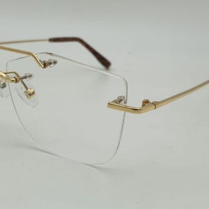May include: Rimless eyeglasses with a gold-colored metal frame and temples. The lenses are clear, and the nose pads are small and gold. The temples have brown tips. The glasses are designed for vision correction.