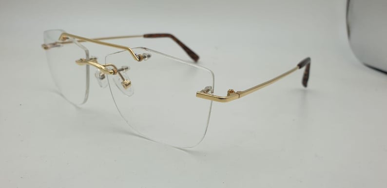 May include: Rimless eyeglasses with a gold-colored metal frame and temples. The lenses are clear, and the nose pads are small and gold. The temples have brown tips. The glasses are designed for vision correction.