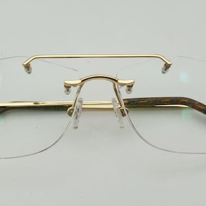 May include: Rimless eyeglasses with a gold-colored metal frame and temples with a brown and green pattern. The glasses have a double bridge and clear lenses. The overall design is modern and stylish.