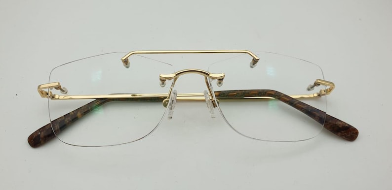 May include: Rimless eyeglasses with a gold-colored metal frame and temples with a brown and green pattern. The glasses have a double bridge and clear lenses. The overall design is modern and stylish.