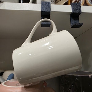 May include: A white ceramic mug hanging from a black mug holder. The mug has a curved handle and a glossy finish. Other mugs are visible in the background, along with a shelf and decorative items.