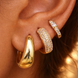 Puede incluir: Primer plano de tres pendientes dorados. Uno es un aro grueso y pulido. El segundo es un aro curvo cubierto de pequeñas piedras brillantes. El tercero es un aro pequeño y recto con tres piedras.