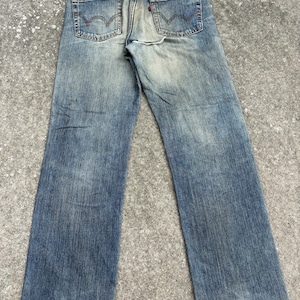 K&ouml;nnte beinhalten: Verblasste blaue Jeans mit einem Used-Look. Die Jeans haben ein klassisches F&uuml;nf-Taschen-Design, einen roten Logoaufn&auml;her und ein abgenutztes Aussehen. Die Jeans haben einen geraden Beinverlauf und sind am Saum leicht ausgefranst.