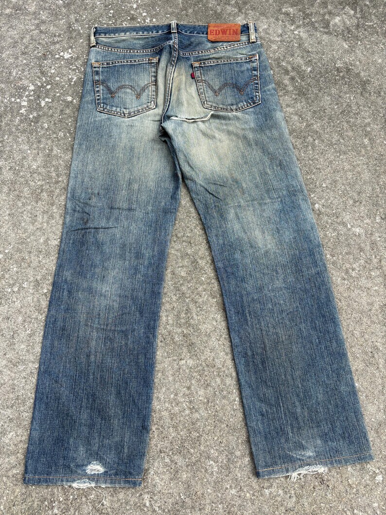 K&ouml;nnte beinhalten: Verblasste blaue Jeans mit einem Used-Look. Die Jeans haben ein klassisches F&uuml;nf-Taschen-Design, einen roten Logoaufn&auml;her und ein abgenutztes Aussehen. Die Jeans haben einen geraden Beinverlauf und sind am Saum leicht ausgefranst.