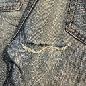 K&ouml;nnte beinhalten: Nahaufnahme einer hellblauen Jeans mit einer ausgefransten Tasche. Die Tasche hat eine ausgefranste &Ouml;ffnung, die die wei&szlig;en F&auml;den darunter freilegt. Die Jeans weist Gebrauchsspuren auf, mit sichtbaren N&auml;hten und einem verblichenen Aussehen.