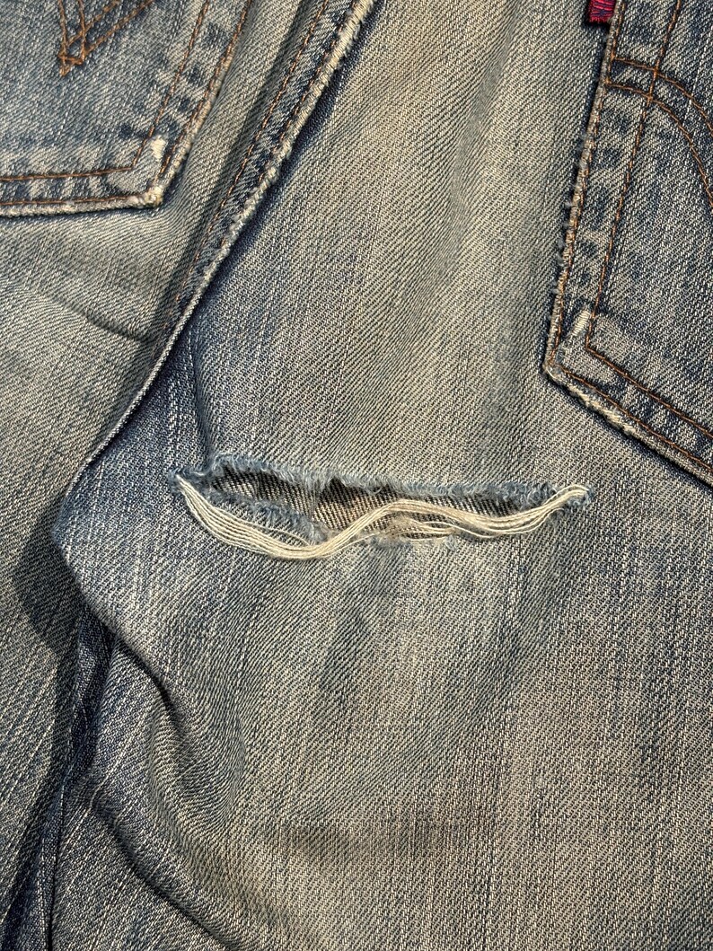 K&ouml;nnte beinhalten: Nahaufnahme einer hellblauen Jeans mit einer ausgefransten Tasche. Die Tasche hat eine ausgefranste &Ouml;ffnung, die die wei&szlig;en F&auml;den darunter freilegt. Die Jeans weist Gebrauchsspuren auf, mit sichtbaren N&auml;hten und einem verblichenen Aussehen.