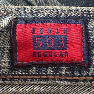 K&ouml;nnte beinhalten: Nahaufnahme eines Jeans-Etiketts. Das rote rechteckige Etikett zeigt den Markennamen "EDWIN" und die Nummer "503" in Marineblau. Das Wort "REGULAR" ist ebenfalls sichtbar. Das Etikett ist auf den blauen Denim-Stoff gen&auml;ht.