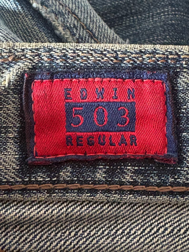 K&ouml;nnte beinhalten: Nahaufnahme eines Jeans-Etiketts. Das rote rechteckige Etikett zeigt den Markennamen "EDWIN" und die Nummer "503" in Marineblau. Das Wort "REGULAR" ist ebenfalls sichtbar. Das Etikett ist auf den blauen Denim-Stoff gen&auml;ht.