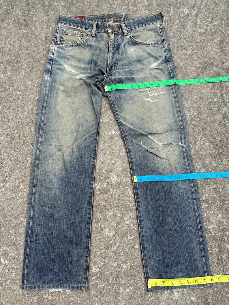 K&ouml;nnte beinhalten: Verblasste blaue Jeans aus Denim mit abgenutzten Details, einschlie&szlig;lich Rissen und Gebrauchsspuren. Die Jeans haben ein klassisches F&uuml;nf-Taschen-Design und einen Knopf- und Rei&szlig;verschluss. Ma&szlig;e in Zentimetern.