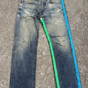 K&ouml;nnte beinhalten: Verblasste blaue Jeans mit abgenutzten Details, einschlie&szlig;lich Rissen und Gebrauchsspuren. Ein gr&uuml;nes Ma&szlig;band zeigt die Bundweite und ein blaues Ma&szlig;band die L&auml;nge. Die Jeans liegen flach auf einer grauen Oberfl&auml;che.