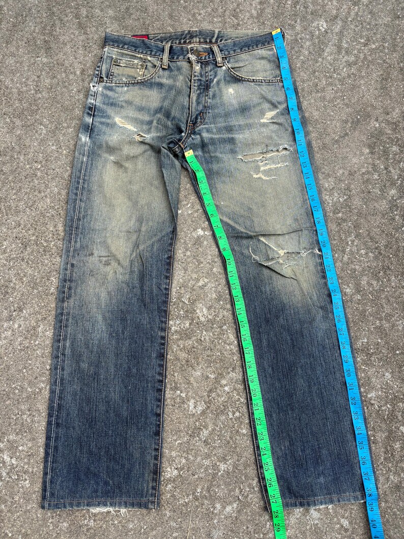 K&ouml;nnte beinhalten: Verblasste blaue Jeans mit abgenutzten Details, einschlie&szlig;lich Rissen und Gebrauchsspuren. Ein gr&uuml;nes Ma&szlig;band zeigt die Bundweite und ein blaues Ma&szlig;band die L&auml;nge. Die Jeans liegen flach auf einer grauen Oberfl&auml;che.
