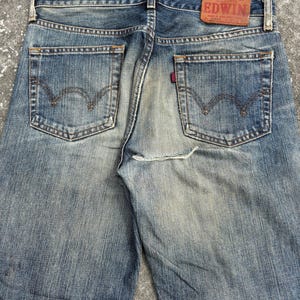 K&ouml;nnte beinhalten: Verblasste blaue Jeans mit klassischem F&uuml;nf-Taschen-Design. Die R&uuml;ckansicht zeigt die Taschen, G&uuml;rtelschlaufen und einen Lederaufn&auml;her mit dem Markennamen "EDWIN". Die Jeans haben einen abgenutzten, Vintage-Look.