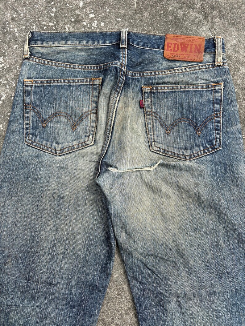 K&ouml;nnte beinhalten: Verblasste blaue Jeans mit klassischem F&uuml;nf-Taschen-Design. Die R&uuml;ckansicht zeigt die Taschen, G&uuml;rtelschlaufen und einen Lederaufn&auml;her mit dem Markennamen "EDWIN". Die Jeans haben einen abgenutzten, Vintage-Look.