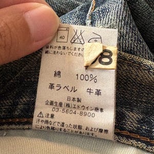 K&ouml;nnte beinhalten: Nahaufnahme eines Denim-Kleidungsst&uuml;cketiketts mit japanischem Text und Pflegesymbolen. Das Etikett gibt an, dass der Stoff aus 100 % Baumwolle besteht und ein Lederetikett hat. Waschanweisungen sind ebenfalls enthalten. Das Etikett ist an einem blauen Denim-Stoff befestigt.