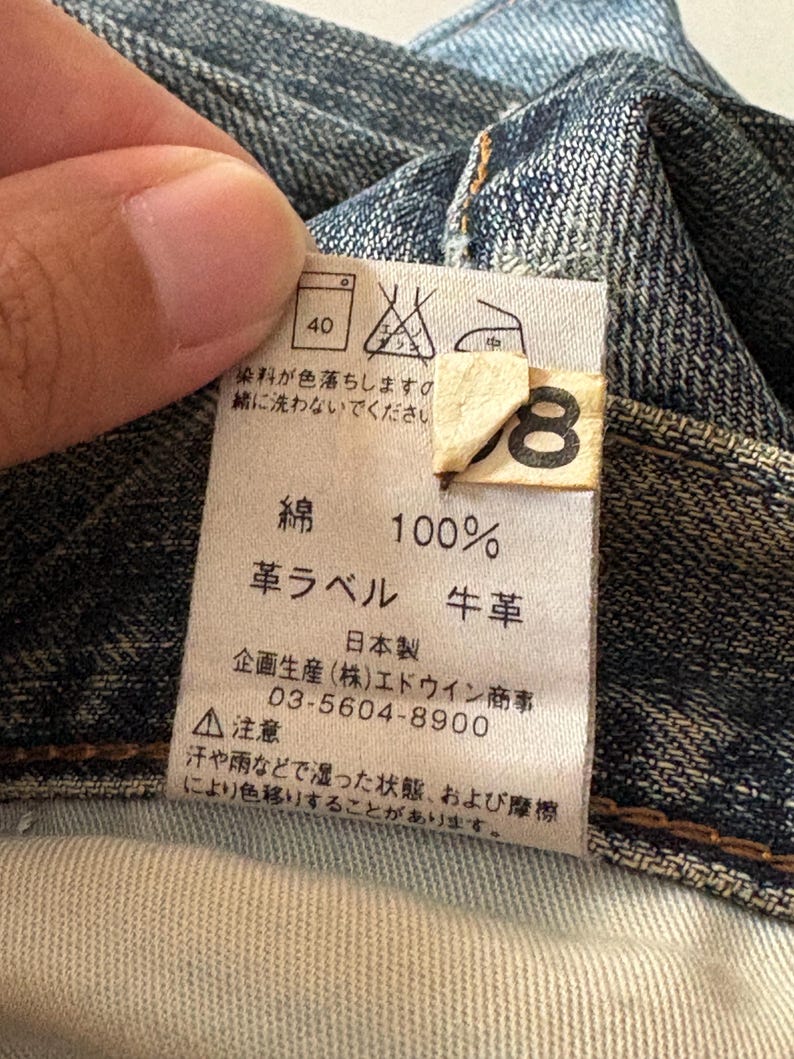 K&ouml;nnte beinhalten: Nahaufnahme eines Denim-Kleidungsst&uuml;cketiketts mit japanischem Text und Pflegesymbolen. Das Etikett gibt an, dass der Stoff aus 100 % Baumwolle besteht und ein Lederetikett hat. Waschanweisungen sind ebenfalls enthalten. Das Etikett ist an einem blauen Denim-Stoff befestigt.