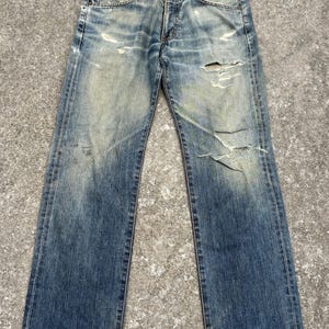 K&ouml;nnte beinhalten: Verblasste blaue Jeans aus Denim mit abgenutzten Details, einschlie&szlig;lich Rissen und Gebrauchsspuren. Die Jeans haben ein klassisches F&uuml;nf-Taschen-Design und einen Knopf- und Rei&szlig;verschluss. Der Denim hat einen abgenutzten Vintage-Look.