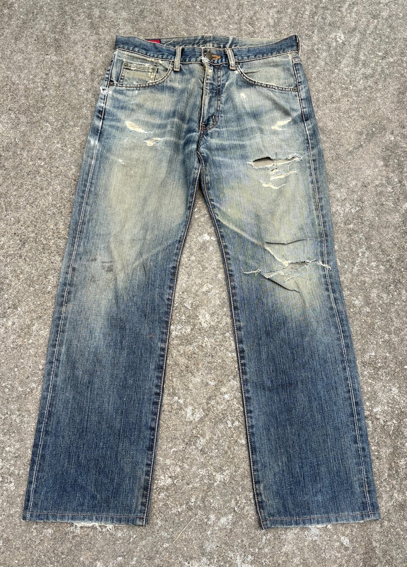K&ouml;nnte beinhalten: Verblasste blaue Jeans aus Denim mit abgenutzten Details, einschlie&szlig;lich Rissen und Gebrauchsspuren. Die Jeans haben ein klassisches F&uuml;nf-Taschen-Design und einen Knopf- und Rei&szlig;verschluss. Der Denim hat einen abgenutzten Vintage-Look.