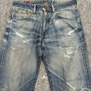 K&ouml;nnte beinhalten: Verblasste blaue Jeans mit Distressed-Details, einschlie&szlig;lich Rissen und abgenutzten Stellen. Die Jeans haben ein klassisches F&uuml;nf-Taschen-Design und einen Knopfverschluss. Der Denim weist Gebrauchsspuren auf und verleiht ihm einen Vintage-Look.