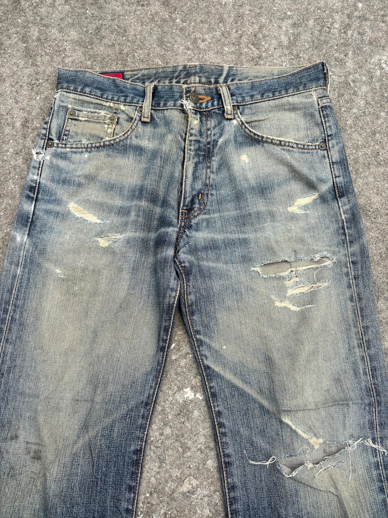 K&ouml;nnte beinhalten: Verblasste blaue Jeans mit Distressed-Details, einschlie&szlig;lich Rissen und abgenutzten Stellen. Die Jeans haben ein klassisches F&uuml;nf-Taschen-Design und einen Knopfverschluss. Der Denim weist Gebrauchsspuren auf und verleiht ihm einen Vintage-Look.