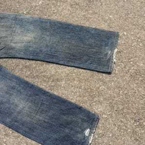 K&ouml;nnte beinhalten: Nahaufnahme einer blauen Jeans aus Denim mit Used-Details. Die Jeans hat eine verwaschene Waschung und sichtbare Gebrauchsspuren. Die S&auml;ume sind ausgefranst, was den Vintage-Look unterstreicht. Die Jeans liegt auf einer strukturierten Oberfl&auml;che.