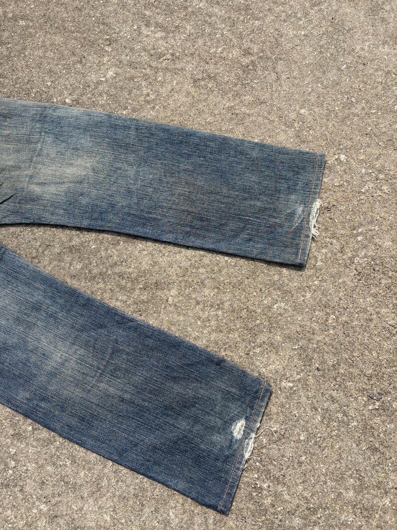 K&ouml;nnte beinhalten: Nahaufnahme einer blauen Jeans aus Denim mit Used-Details. Die Jeans hat eine verwaschene Waschung und sichtbare Gebrauchsspuren. Die S&auml;ume sind ausgefranst, was den Vintage-Look unterstreicht. Die Jeans liegt auf einer strukturierten Oberfl&auml;che.