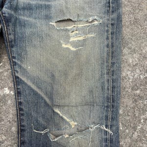K&ouml;nnte beinhalten: Nahaufnahme einer Jeans aus blauem Denim mit Used-Look. Die Jeans hat mehrere Risse und ausgefranste Kanten mit sichtbaren Abnutzungserscheinungen und Ausbleichen. Der Stoff zeigt eine Mischung aus hellen und dunklen Blaut&ouml;nen, was einen Vintage-Look erzeugt. Die Jeans ist ein klassischer Stil.