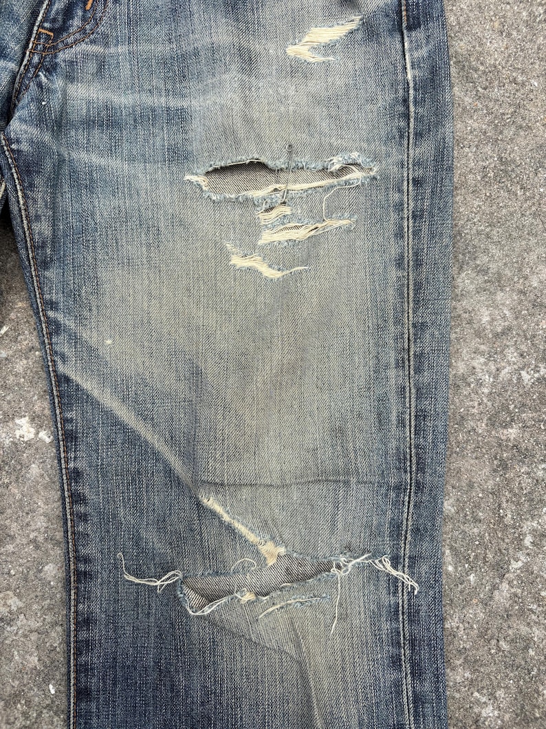 K&ouml;nnte beinhalten: Nahaufnahme einer Jeans aus blauem Denim mit Used-Look. Die Jeans hat mehrere Risse und ausgefranste Kanten mit sichtbaren Abnutzungserscheinungen und Ausbleichen. Der Stoff zeigt eine Mischung aus hellen und dunklen Blaut&ouml;nen, was einen Vintage-Look erzeugt. Die Jeans ist ein klassischer Stil.