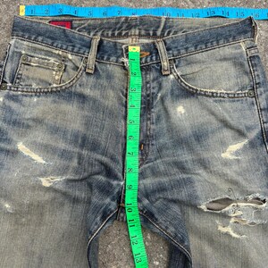 K&ouml;nnte beinhalten: Nahaufnahme einer Jeans aus blauem Denim mit Used-Look und zerrissenen Details. Ein blaues Ma&szlig;band misst die Taille, und ein gr&uuml;nes Ma&szlig;band zeigt die Innenbeinl&auml;nge. Die Jeans hat etwa 40 cm Taillenweite.