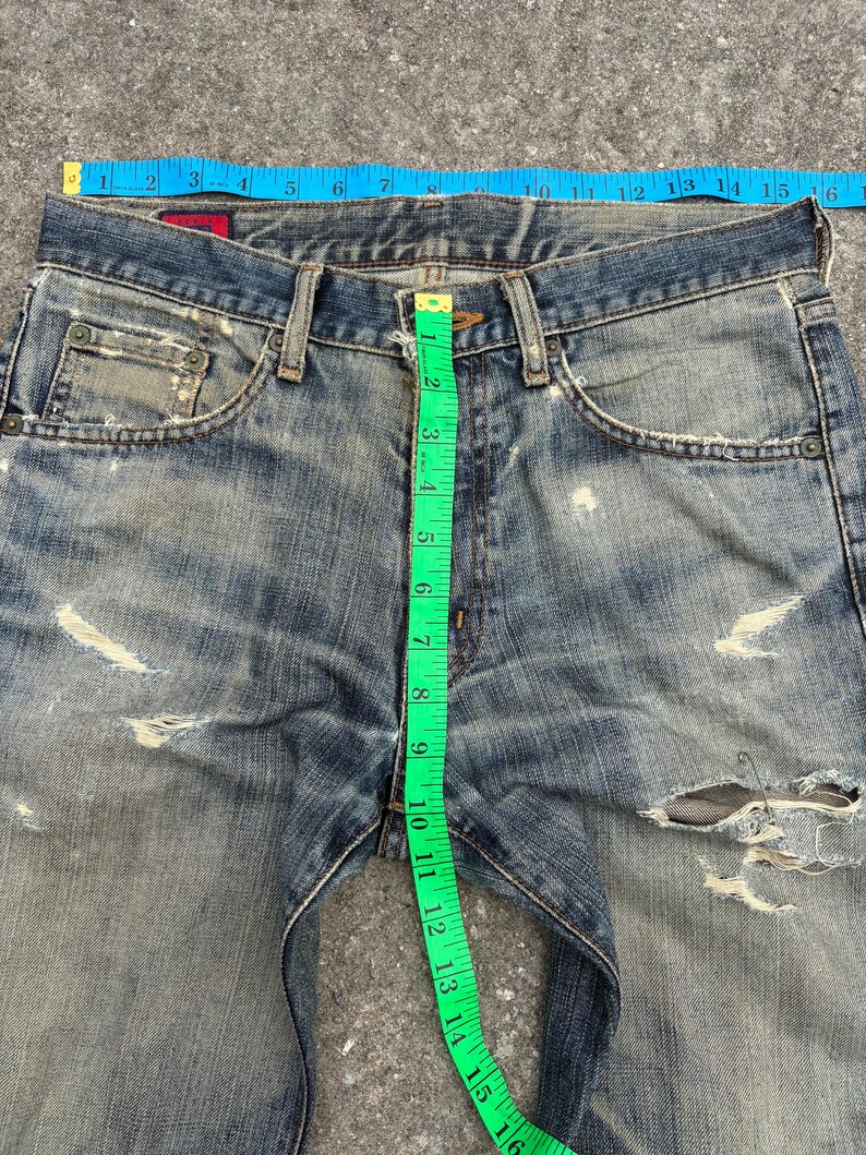 K&ouml;nnte beinhalten: Nahaufnahme einer Jeans aus blauem Denim mit Used-Look und zerrissenen Details. Ein blaues Ma&szlig;band misst die Taille, und ein gr&uuml;nes Ma&szlig;band zeigt die Innenbeinl&auml;nge. Die Jeans hat etwa 40 cm Taillenweite.