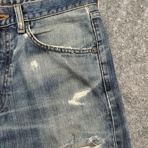K&ouml;nnte beinhalten: Nahaufnahme einer blauen Jeans mit Used-Details. Die Jeans hat ein klassisches F&uuml;nf-Taschen-Design, einen Knopf- und Rei&szlig;verschluss sowie sichtbare Abnutzungserscheinungen, einschlie&szlig;lich Rissen.