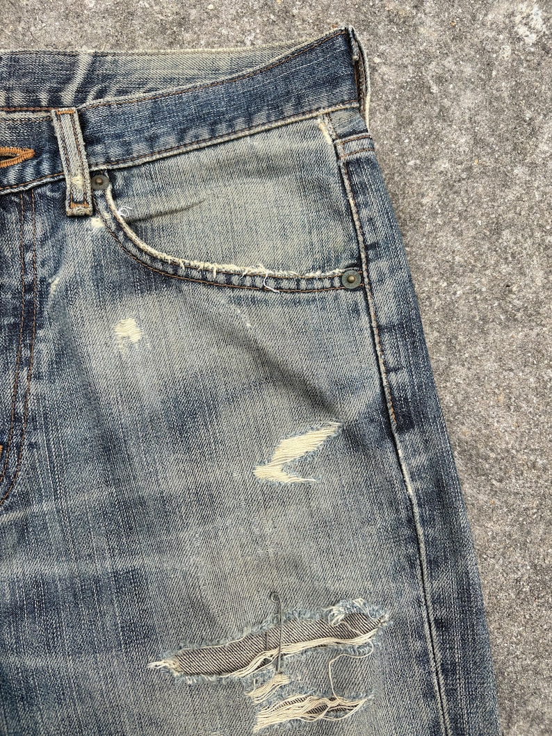 K&ouml;nnte beinhalten: Nahaufnahme einer blauen Jeans mit Used-Details. Die Jeans hat ein klassisches F&uuml;nf-Taschen-Design, einen Knopf- und Rei&szlig;verschluss sowie sichtbare Abnutzungserscheinungen, einschlie&szlig;lich Rissen.