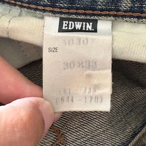 K&ouml;nnte beinhalten: Nahaufnahme eines Jeanskleidungsst&uuml;cks mit Etikett. Das Etikett lautet "EDWIN" in fetten, schwarzen Buchstaben. Unter dem Markennamen gibt das Etikett eine Gr&ouml;&szlig;e von 30x30 und 30x33 an. Zus&auml;tzliche Nummern sind aufgedruckt.