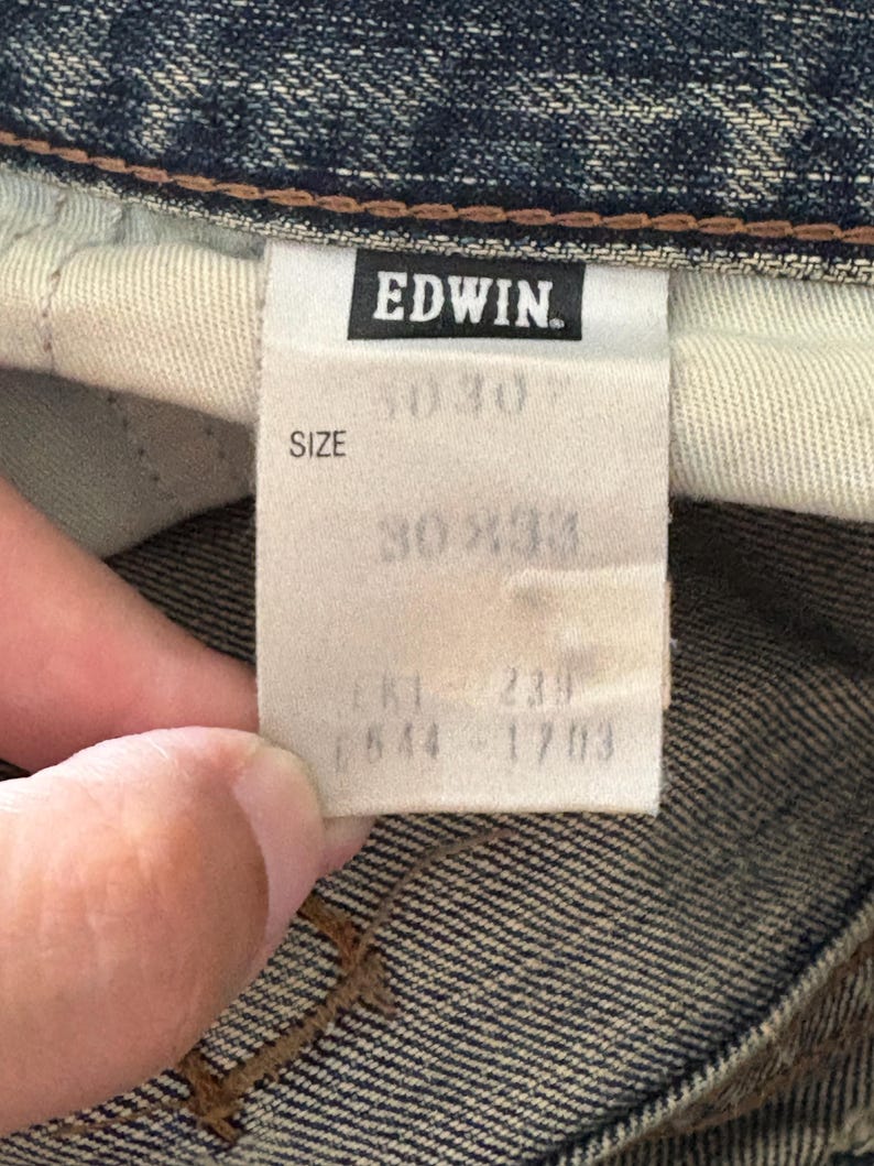 K&ouml;nnte beinhalten: Nahaufnahme eines Jeanskleidungsst&uuml;cks mit Etikett. Das Etikett lautet "EDWIN" in fetten, schwarzen Buchstaben. Unter dem Markennamen gibt das Etikett eine Gr&ouml;&szlig;e von 30x30 und 30x33 an. Zus&auml;tzliche Nummern sind aufgedruckt.