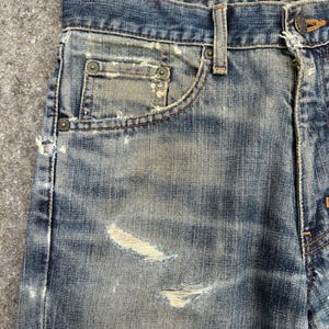 K&ouml;nnte beinhalten: Nahaufnahme einer Jeans aus blauem Denim mit Used-Look. Die Jeans hat eine verblasste Waschung, sichtbare Abnutzung und mehrere besch&auml;digte Bereiche. Die Kanten der Taschen und die N&auml;hte weisen Gebrauchsspuren auf. Die Jeans hat einen Knopfverschluss und G&uuml;rtelschlaufen.