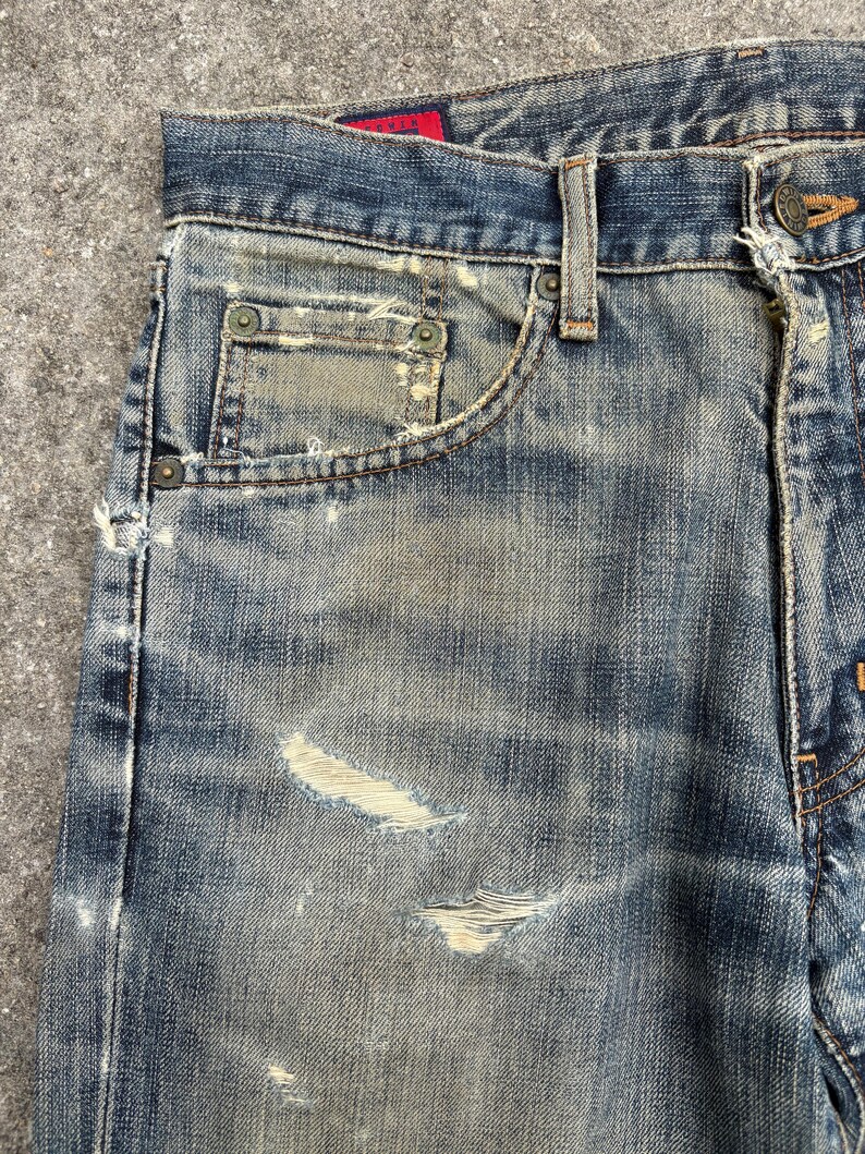 K&ouml;nnte beinhalten: Nahaufnahme einer Jeans aus blauem Denim mit Used-Look. Die Jeans hat eine verblasste Waschung, sichtbare Abnutzung und mehrere besch&auml;digte Bereiche. Die Kanten der Taschen und die N&auml;hte weisen Gebrauchsspuren auf. Die Jeans hat einen Knopfverschluss und G&uuml;rtelschlaufen.