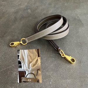 May include: A taupe leather bag strap featuring a textured surface, white contrast stitching, and gold-tone metal lobster claw clasps at both ends. An inset shows the strap attached to a taupe handbag worn with a white cable knit sweater.