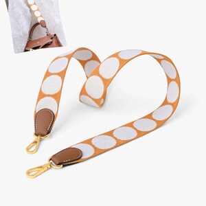 May include: A wide, woven bag strap featuring a mustard yellow background with large, white circular polka dots. The strap ends are finished with brown leather tabs and gold-toned metal lobster claw clasps for attachment to a handbag.