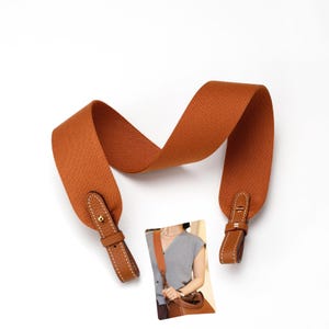 May include: A wide, textured, burnt orange fabric bag strap featuring leather end tabs with white contrast stitching and gold-toned metal hardware. The strap is shown in a curved position, accompanied by a small inset photo of the strap in use.